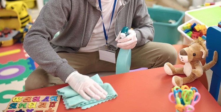 Junger glatzköpfiger Mann, Vorschullehrer mit medizinischer Maske, reinigt den Tisch im Kindergarten